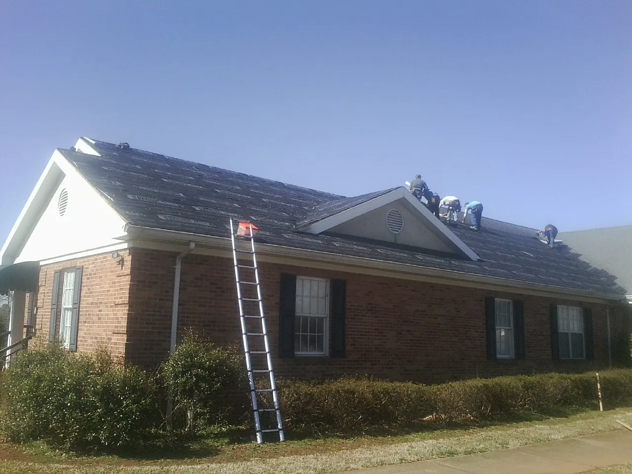 Crew tearing shingles off a brick ranch roof with a ladder set — Emergency Roof Repair work by our crew in Sacred Heart