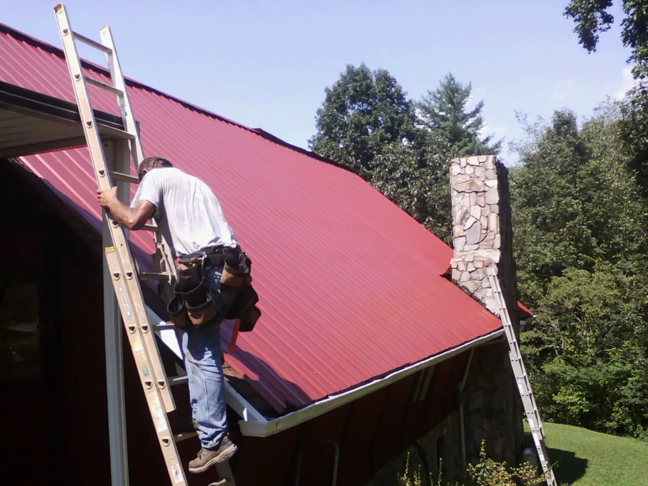 Roofer on a ladder beside a red metal roof and stone chimney — Soffit & Fascia Repair by Roofing Brothers in Sacred Heart